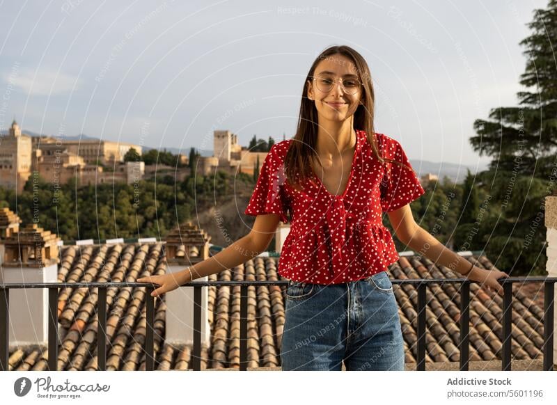 Happy tourist standing by railing against historic Alcazaba while enjoying vacation in Granada, Spain woman eyeglasses smile casual attire tree history alcazaba