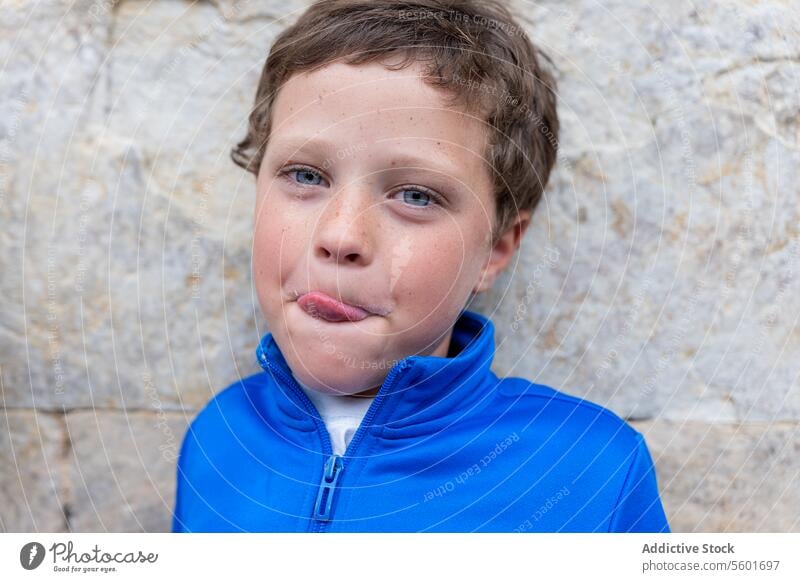 Blue eyed boy looking at the camera with a funny face Boy child humor playful blue jacket stone wall expression kid mischievous childhood cheeky outdoor