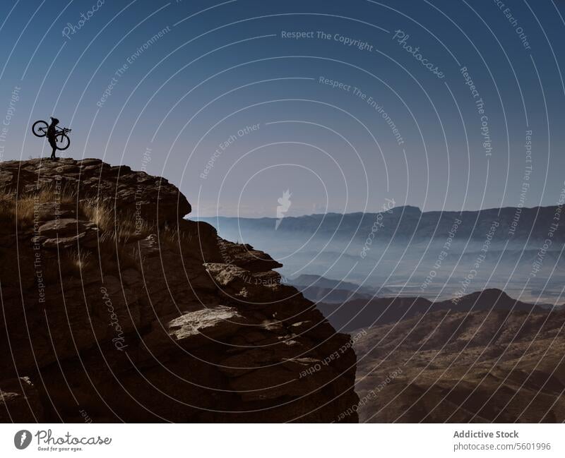 Cyclist on mountain cliff holding a bicycle while admiring the landscape cyclist overlook valley dusk rugged terrain adventure outdoor sport bike cycling