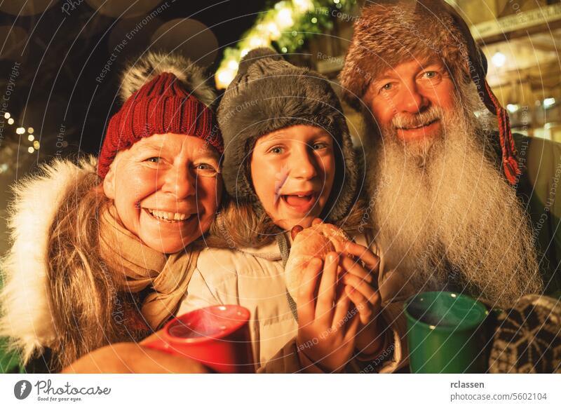 Happy Grandparents with grandchild eating a sausage sandwich and holding mugs of mulled wine at a Christmas market merry christmas gloves traditional