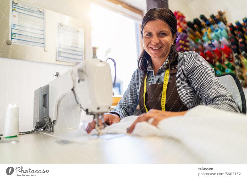 cheerful needlewoman woman with a tape measure around her neck is sitting by a sewing machine in a brightly lit workshop, holding a white silk fabric with colorful spools of thread in the background