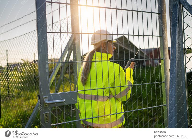 Female technician with walkie-talkie walking towards solar field farm gate technology plant industry electricity worker safety engineer ecological solar park