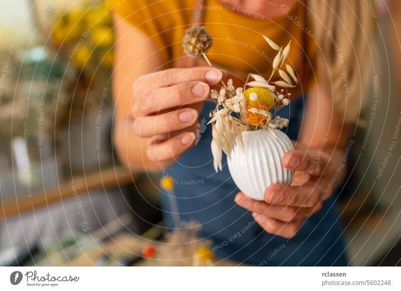 Close-up of hands arranging dried flowers in a small white ribbed vase, blurred background white vase close-up floristry crafting yellow top blue apron diy