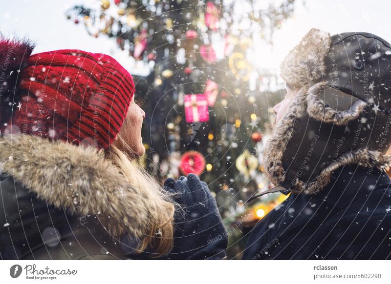 Woman in red beanie and man in fur hat face looking up do a christmas tree at christmas market, snowflakes falling on a winter day, with copyspace for your individual text.