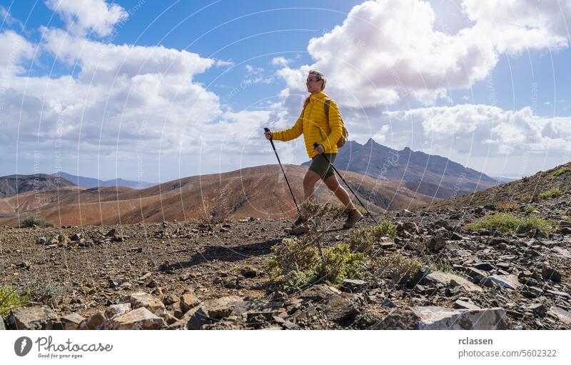 Hiker in yellow jacket walking on mountain trail with cloudy sky and peak in distance fuerteventura hiker hiking outdoor adventure trekking nature wilderness