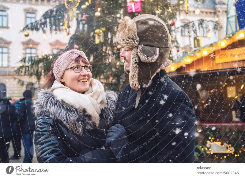 smiling couple in winter clothes stand close at a snowy Christmas market, with festive lights behind them. happy couple hugging friends mulled wine boyfriend