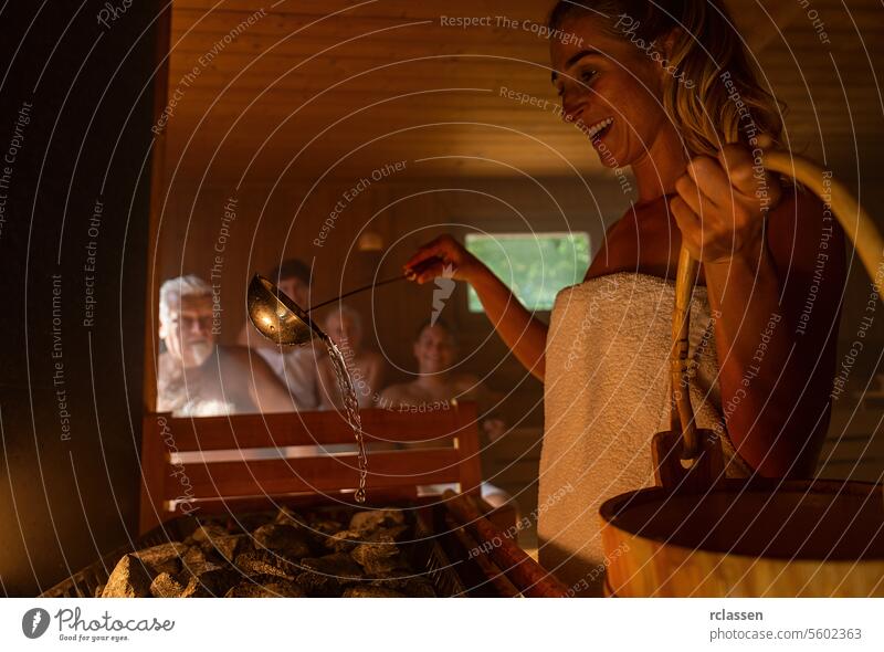 Happy woman pouring water onto hot stone in sauna room with a group of people. Steam an water on the stones, spa and wellness concept, relax in hot finnish sauna.