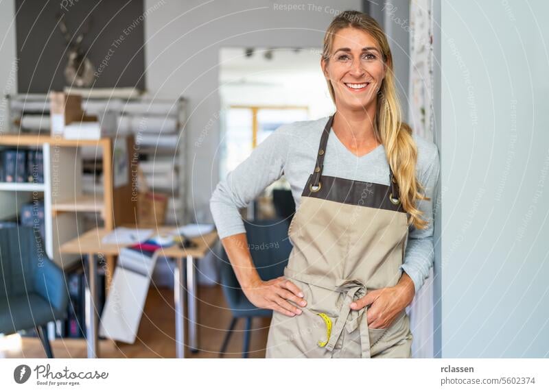 cheerful blonde woman in a gray top and beige apron stands in a tailor workspace. She has a measuring tape and a scissors in her apron pocket and is surrounded by organized craft materials