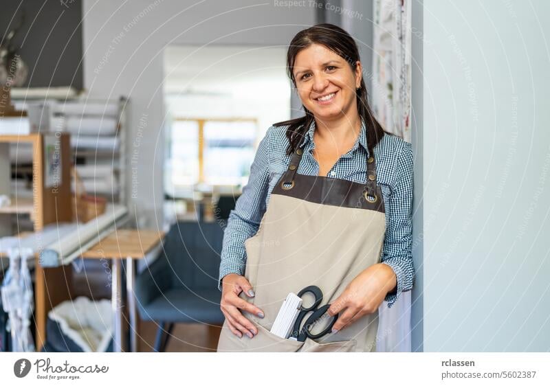 smiling tailor woman in a checkered shirt and beige apron stands in a workspace, holding a pair of scissors and a scale ruler in her apron pocket curtains