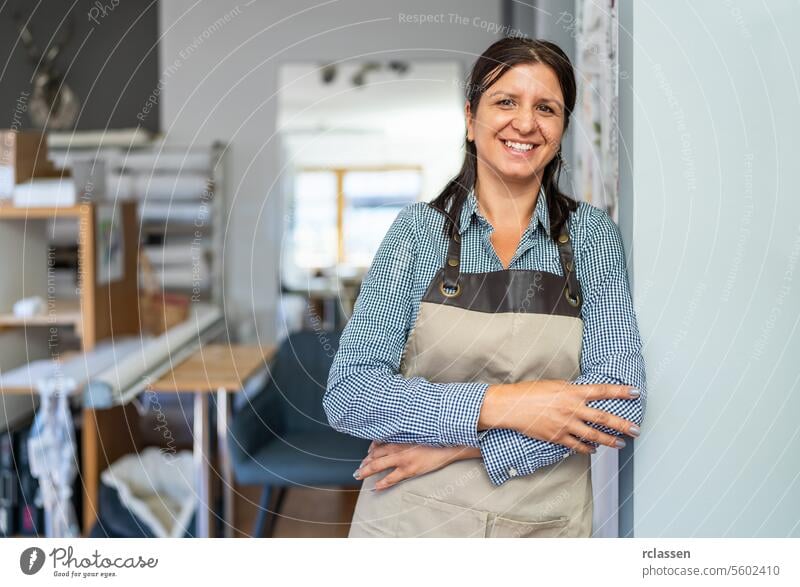 confident woman wearing a checkered shirt and beige apron stands in a workshop with folded arms, smiling towards the camera. scissors curtains profession