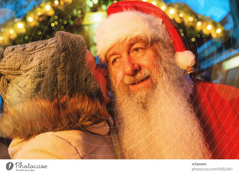 Child whispering to Santa Claus at a Christmas market with festive lights in the background bokeh joyful merry christmas gloves traditional mulled wine