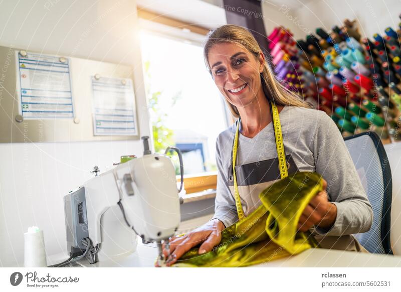 cheerful seamstress woman with a tape measure around her neck is sitting by a sewing machine in a brightly lit workshop, holding a golden silk fabric with colorful spools of thread in the background