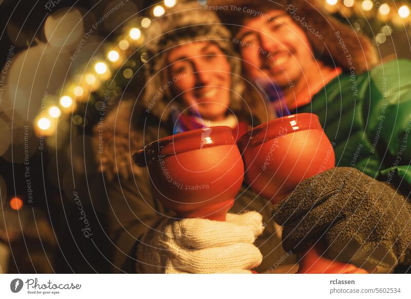 Smiling couple at a Christmas market, holding red mugs with blue flames from fire tongs punch, festive lights behind. tourist mulled wine christmas market