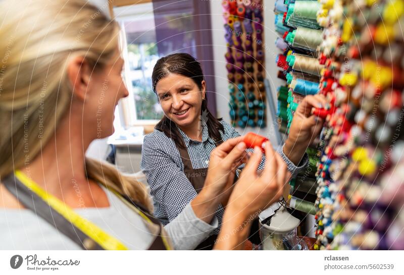 Work colleagues in a tailor shop, one in focus, share a moment in a sewing workspace. They're surrounded by colorful threads, with one holding a spool of red thread