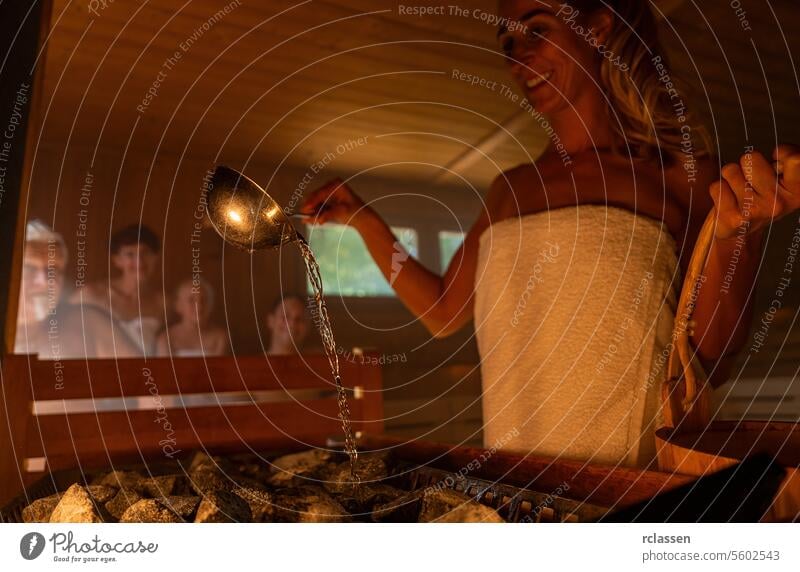 Happy woman holding bucket and spoon pouring water onto hot stone in sauna room with a group of people. Steam an water on the stones, spa and wellness concept, relax in hot finnish sauna.