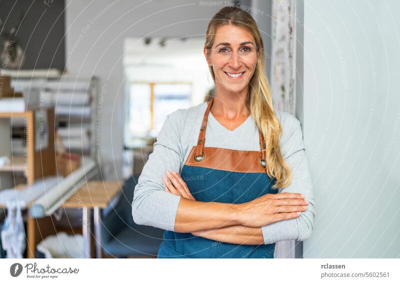 cheerful blonde woman with arms crossed in apron stands  in a tailor workspace. She has a measuring tape and a scissors in her apron pocket and is surrounded by organized craft materials