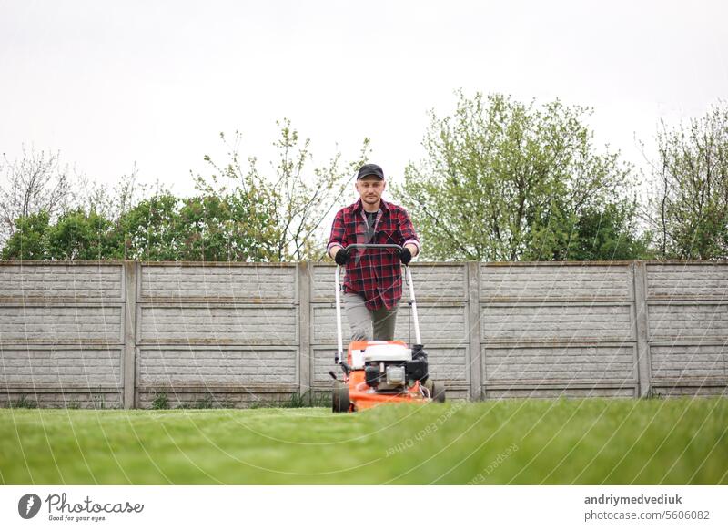 view of man in casual clothes mows lawn with lawn mower at backyard of his house. Husband takes care of garden on spring cloudy day. Modern gasoline garden equipment. Landscaping work