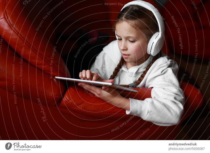 Concentrated teen girl in headphones plays game, communicate on internet social media, listens to music or online studies at home on digital tablet sitting on sofa. Children tech addiction concept