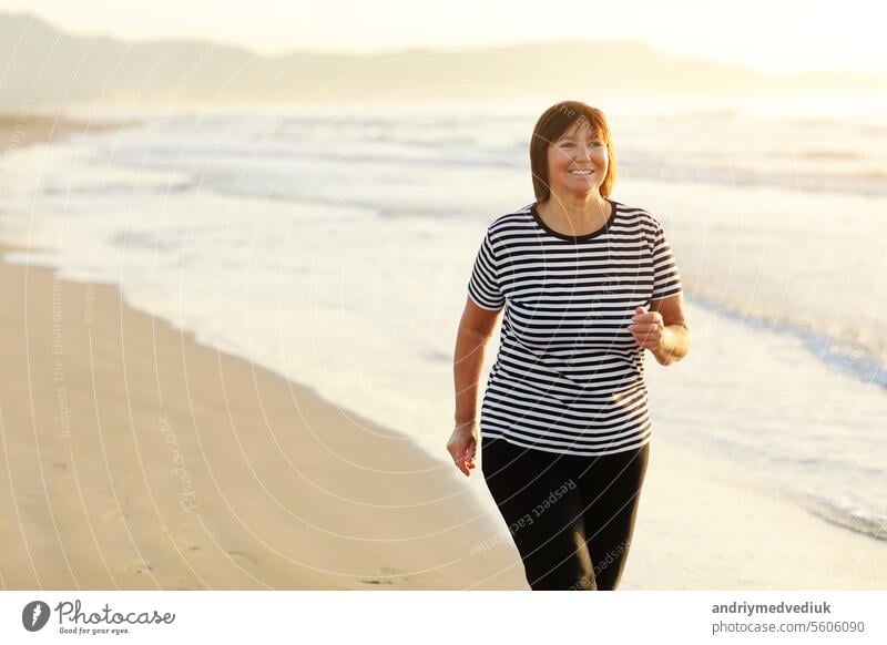 Smiling middle aged woman running on the beach on sunrise. 40s or 50s attractive mature lady in sports clothes doing jogging workout enjoying fitness and healthy lifestyle at beautiful sea landscape.