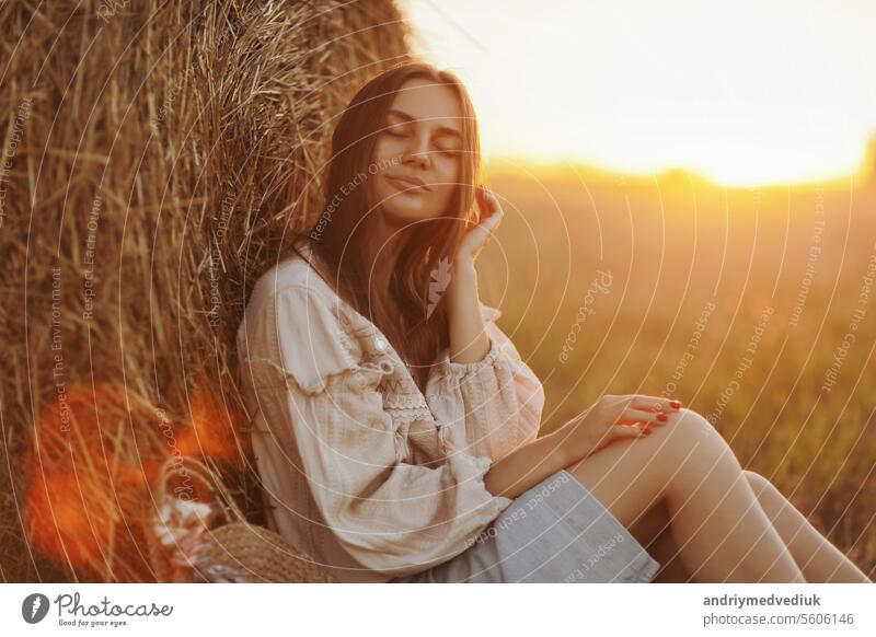 young woman in the beautiful light of the summer sunset in a field is sitting near the straw bales. beautiful romantic girl with long hair outdoors in field