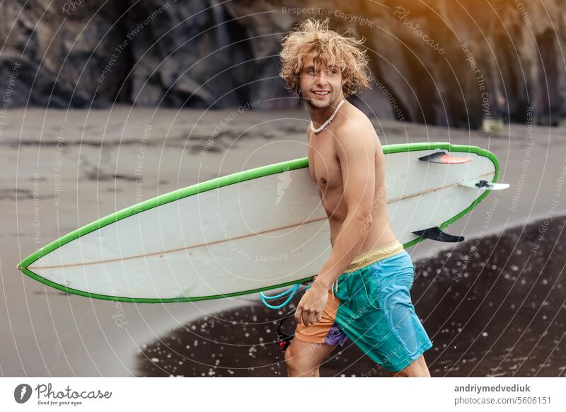 Fit young surfer man with curly blond hair with surfboard goes by the ocean having fun doing extreme water sports, surfing. Travel and healthy lifestyle concept. Sports travel destination.