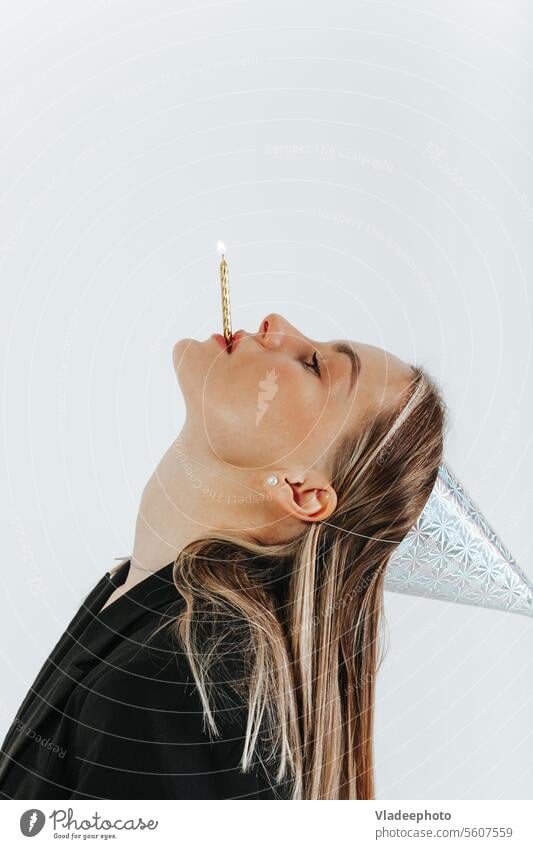 Blonde girl in a festive cap throw back her head and holds a burning birthday candle with her lips mouth woman black light background party hat celebration