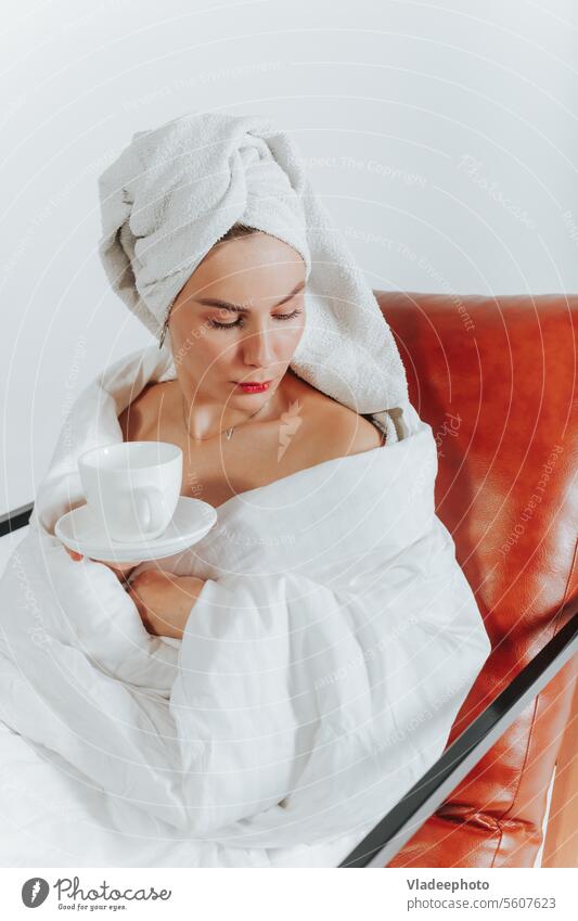 Woman drinking coffee or tea wrapped in a white blanket and towel around head. White background woman morning home lifestyle bathrobe bedding girl lady cozy