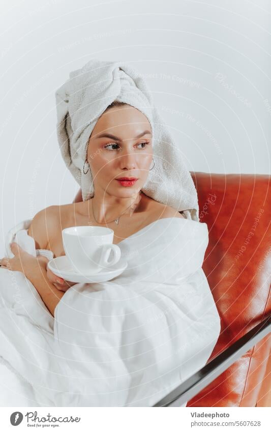 Woman drinking coffee or tea wrapped in a white blanket and towel around head. White background woman morning home lifestyle bathrobe bedding girl lady cozy