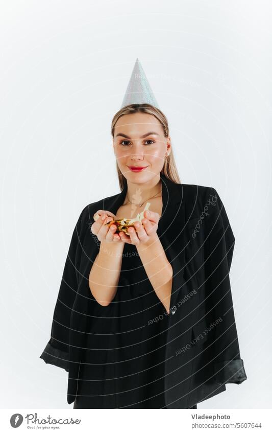 Smiling lady with party hat and holding confetti in hands and looking at camera. White background happy holiday birthday festive white girl isolated whistle