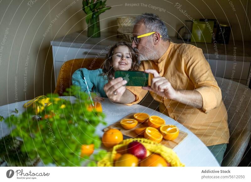 High angle view of senior grandfather kissing on happy granddaughter's forehead while taking selfie over smart phone at dining table at home girl man smartphone