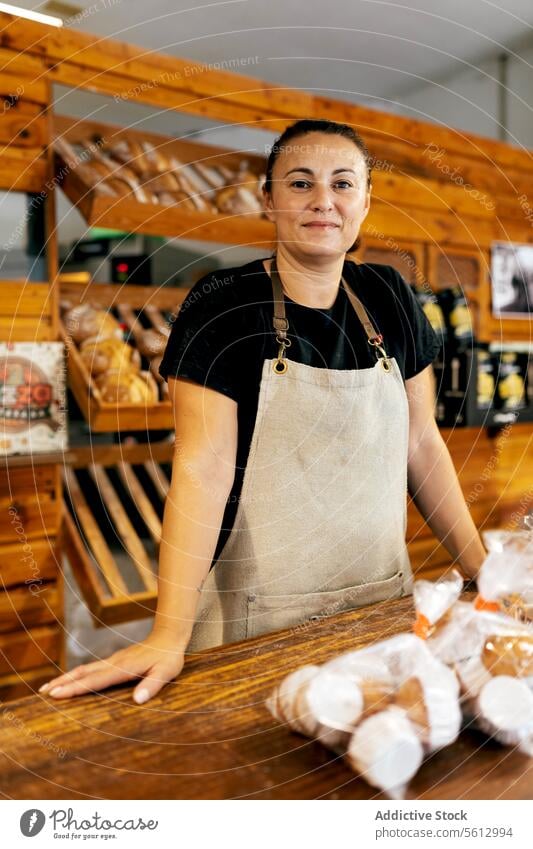 Happy saleswoman standing by bread in bakery shop seller shelf loaf confident smile apron fresh baked food wooden counter positive manufacture production