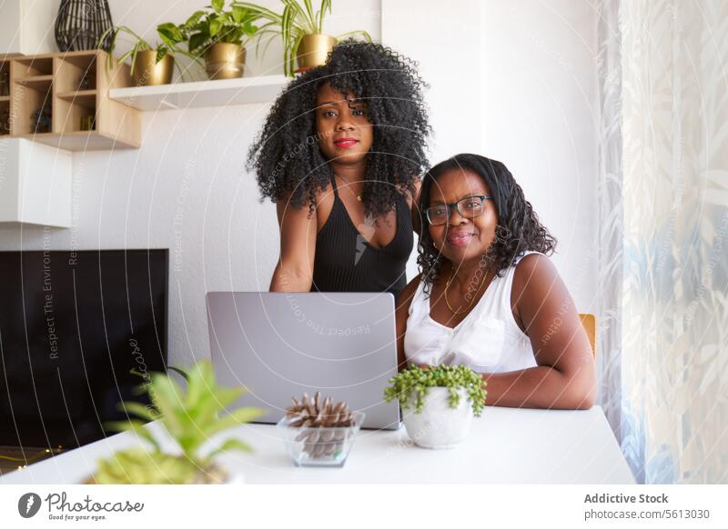 Smiling woman with mother using gadget women daughter happy laptop table afro lifestyle together sitting plant home smiling eyeglasses love domestic curly hair