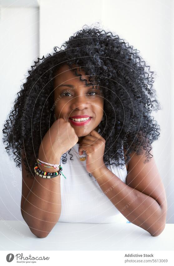 Woman with afro hairstyle at home woman smiling looking at camera touch chin leaning portrait white table black curly lifestyle contemplating domestic lady