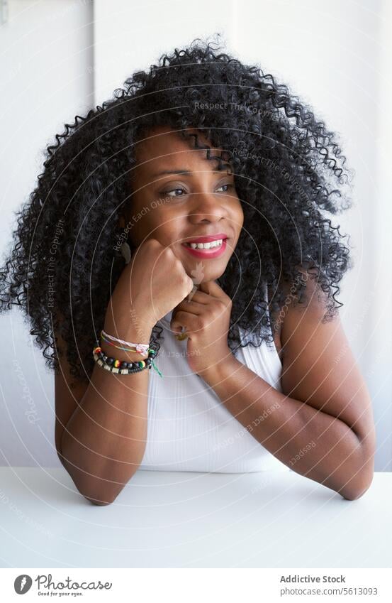 Woman with afro hairstyle at home woman smiling looking away touch chin leaning portrait white table black curly lifestyle contemplating domestic lady apartment