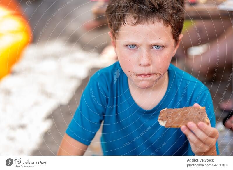 Cute hungry boy dressed in casuals clothes looking at camera while eating sweet chocolate spread with bread for breakfast while sitting in restaurant sandwich