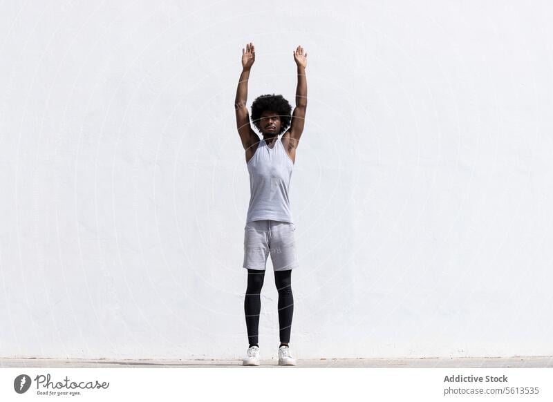 Full body of confident African American male athlete in activewear with concentration stretching arms while standing looking away against white background man