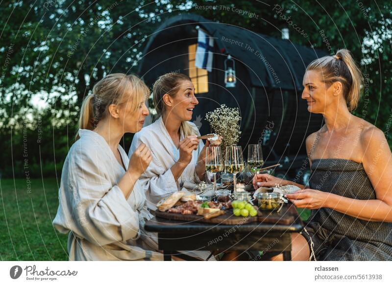 Friends in bathrobes enjoying a meal and wine near a Finnish sauna barrel outdoors fog flag friends finnish sauna enjoyment dining relaxation wellness nature