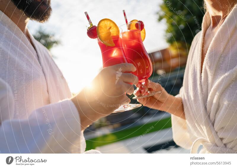 Two people in white bathrobes toasting with red cocktails in the sunlight, outdoors at spa wellness hotel white robes cheers relaxation leisure poolside summer