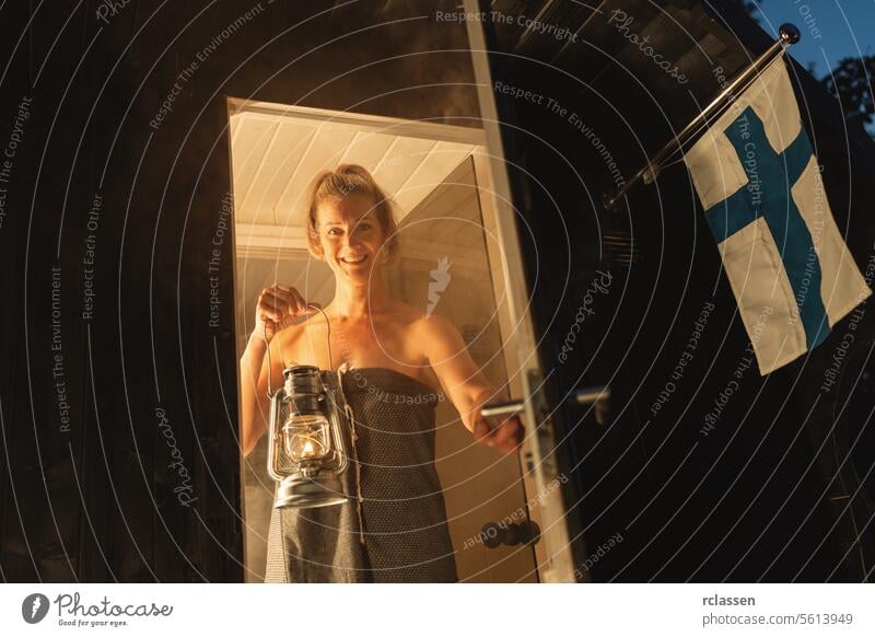 Smiling woman in a towel holding a lantern at the entrance of a Finnish mobile barrel sauna at dusk. finnish barrel sauna smiling woman holding lantern