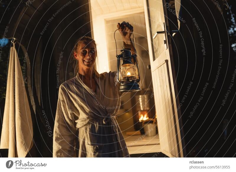 Smiling woman holding a lantern at the entrance of a Finnish barrel sauna during the evening. finnish barrel sauna smiling relaxation health spa wellness