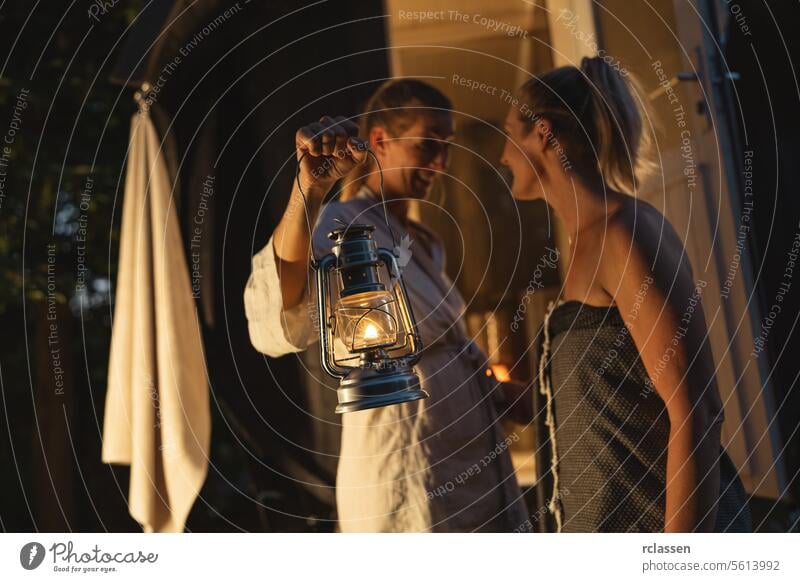 Two women with a lantern by a Finnish barrel sauna at dusk, about to enter. hotel mobile wellness spa finnish barrel sauna entering sauna relaxation wellbeing