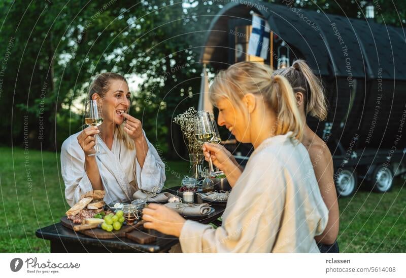 Friends enjoying snacks and wine near a sauna barrel in a lush outdoor setting woman fog friends enjoyment relaxation wellness nature wine glasses spa health