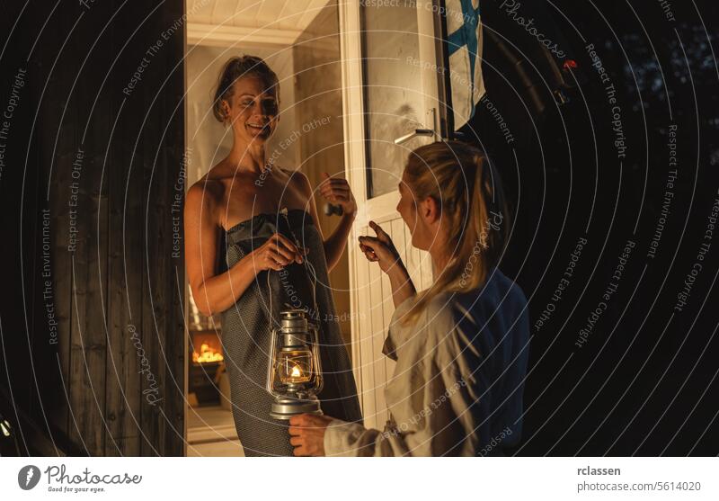 Two women at the entrance of a Finnish mobile barrel sauna, one holding a lantern, smiling and chatting in a cozy atmosphere. pointing finnish sauna friends