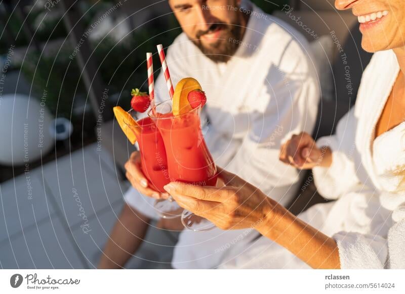 Couple in white robes toasting with fruit garnished cocktails in sunlight at spa hotel bathrobe wellness spa wellness resort couple red cocktails joyful