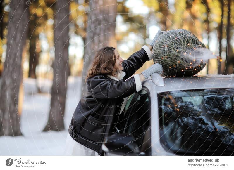 Smiling caucasian woman in winter coat and mittens near car with illuminated Christmas tree on a rooftop on nature in snowy forest. Concept of celebrating New Year holidays. Idea of Xmas mood and fun