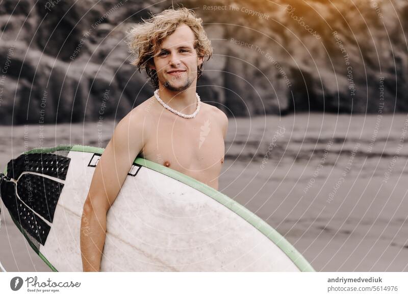 Handsome fit young blond man with mock up surfboard waits for wave to surf spot at sea ocean beach with black sand and looks at camera. Concept of sport, fitness, freedom, happiness, new modern life