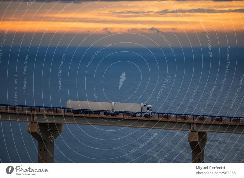 Mega-trailer truck driving over a bridge with the sea and a dramatic sunrise sky on the horizon. mega-trailer sunset transport road-train highway dramatic sky