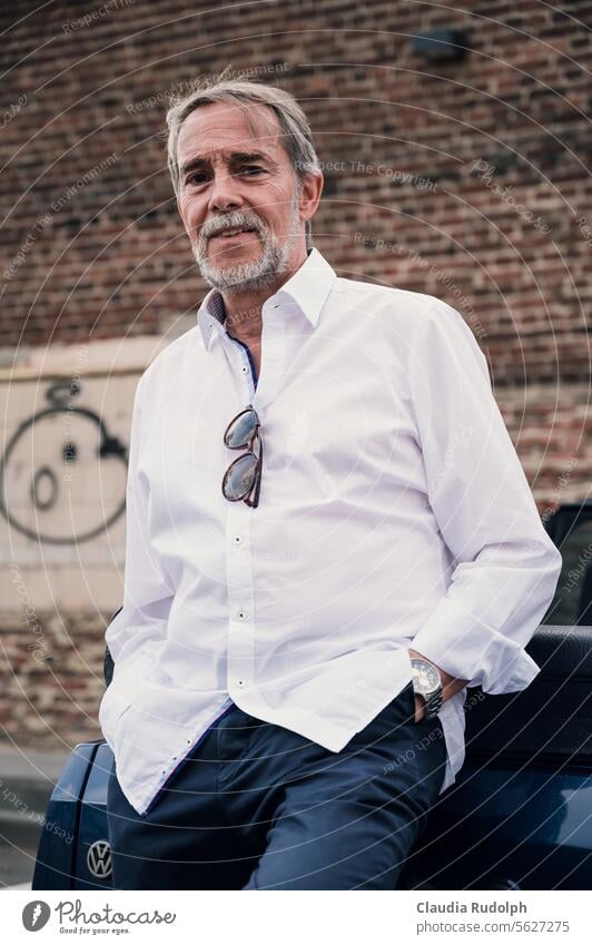 Smiling older gentleman in a white shirt casually leaning against his car, parked in front of an old brick wall with graffiti Best Ager elder mr Man smiling man