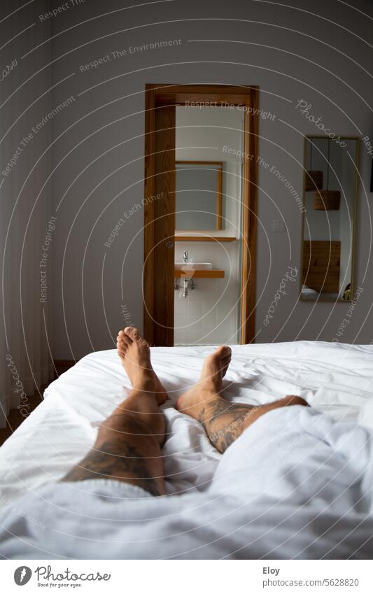 man resting in a hotel room, close-up of the legs of a tattooed man lying on the bed with a white towel, in the background you can see a bathroom and a mirror