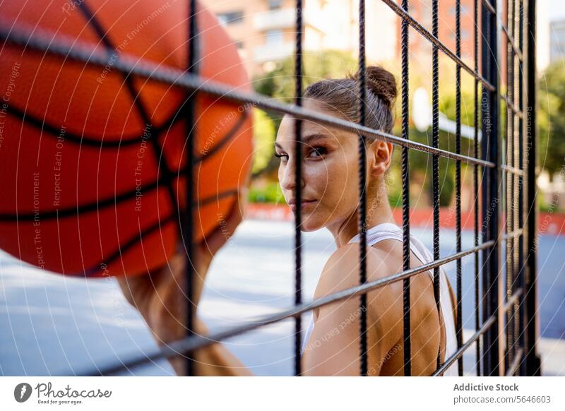 Confident young female athlete in sportswear holding basketball looking at camera seen through fence of playground Sportswoman Basketball Ball Athlete Player